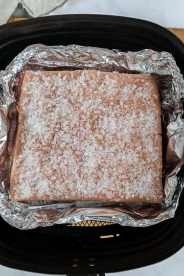 pork belly in an air fryer basket. The top of the pork belly skin is covered with coarse sea salt.